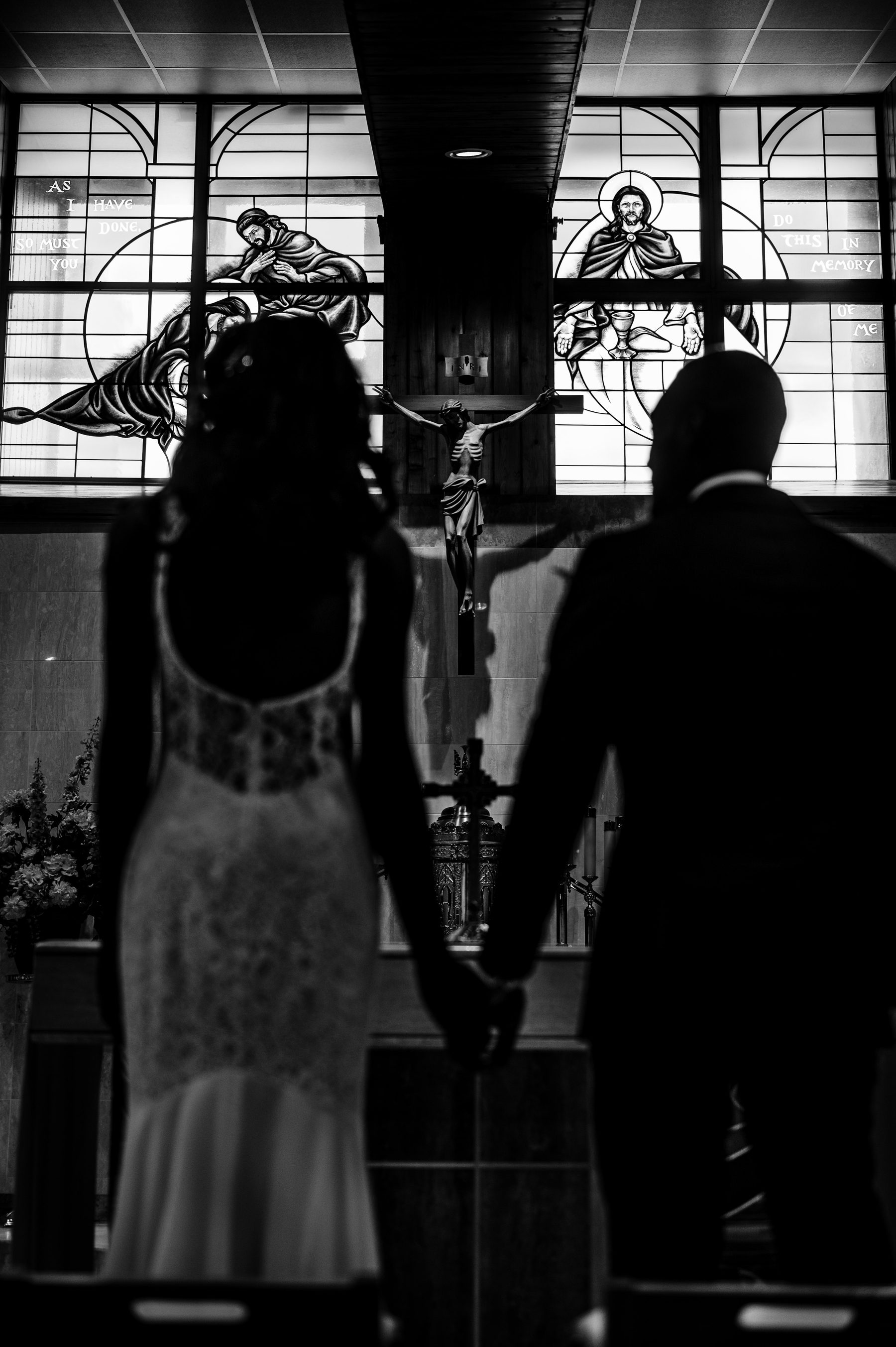 Black and white silhouette of a bride and groom facing a crucifix and stained glass windows