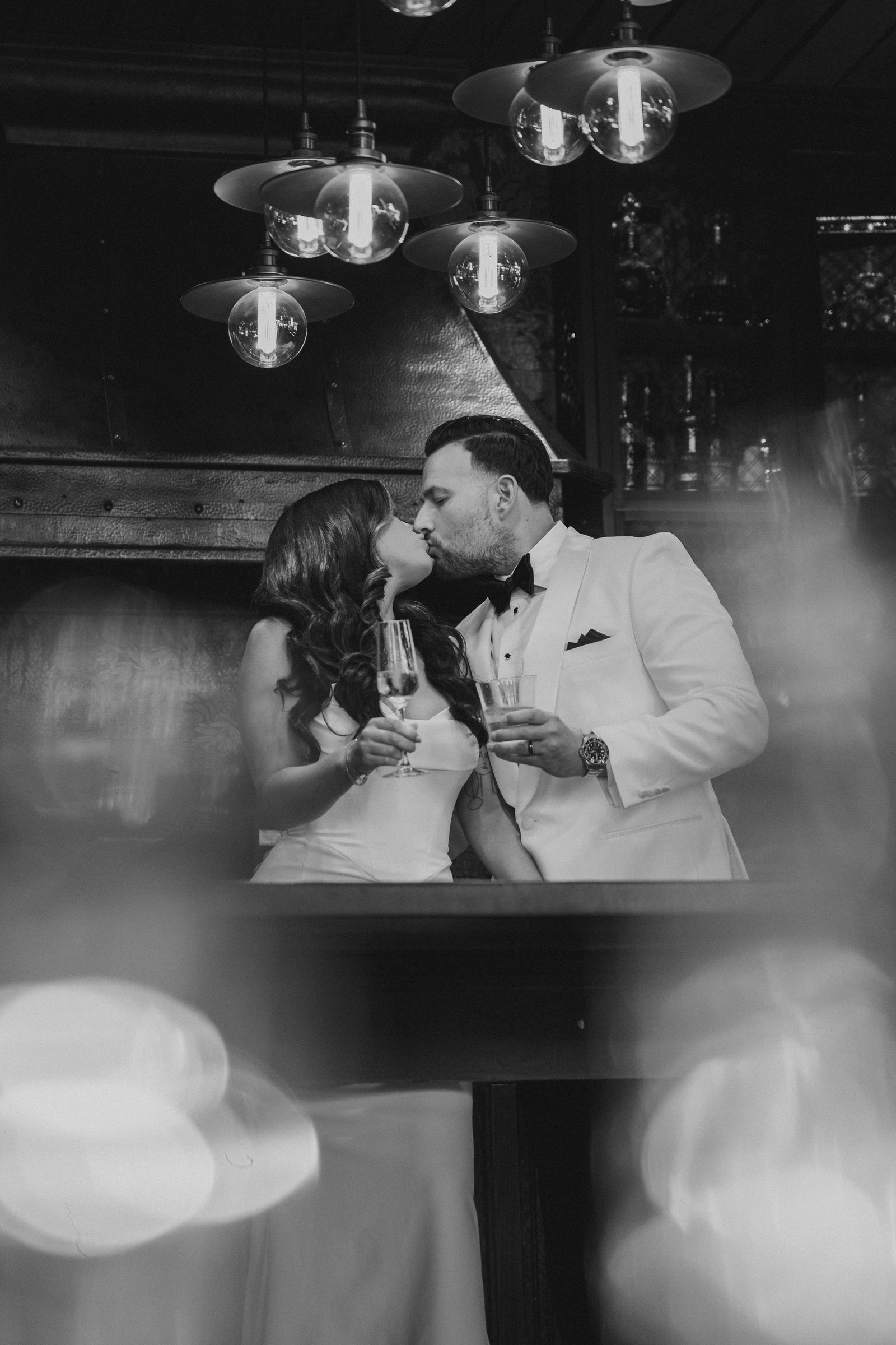 Black and white photo of a bride and groom toasting champagne glasses at a moody bar
