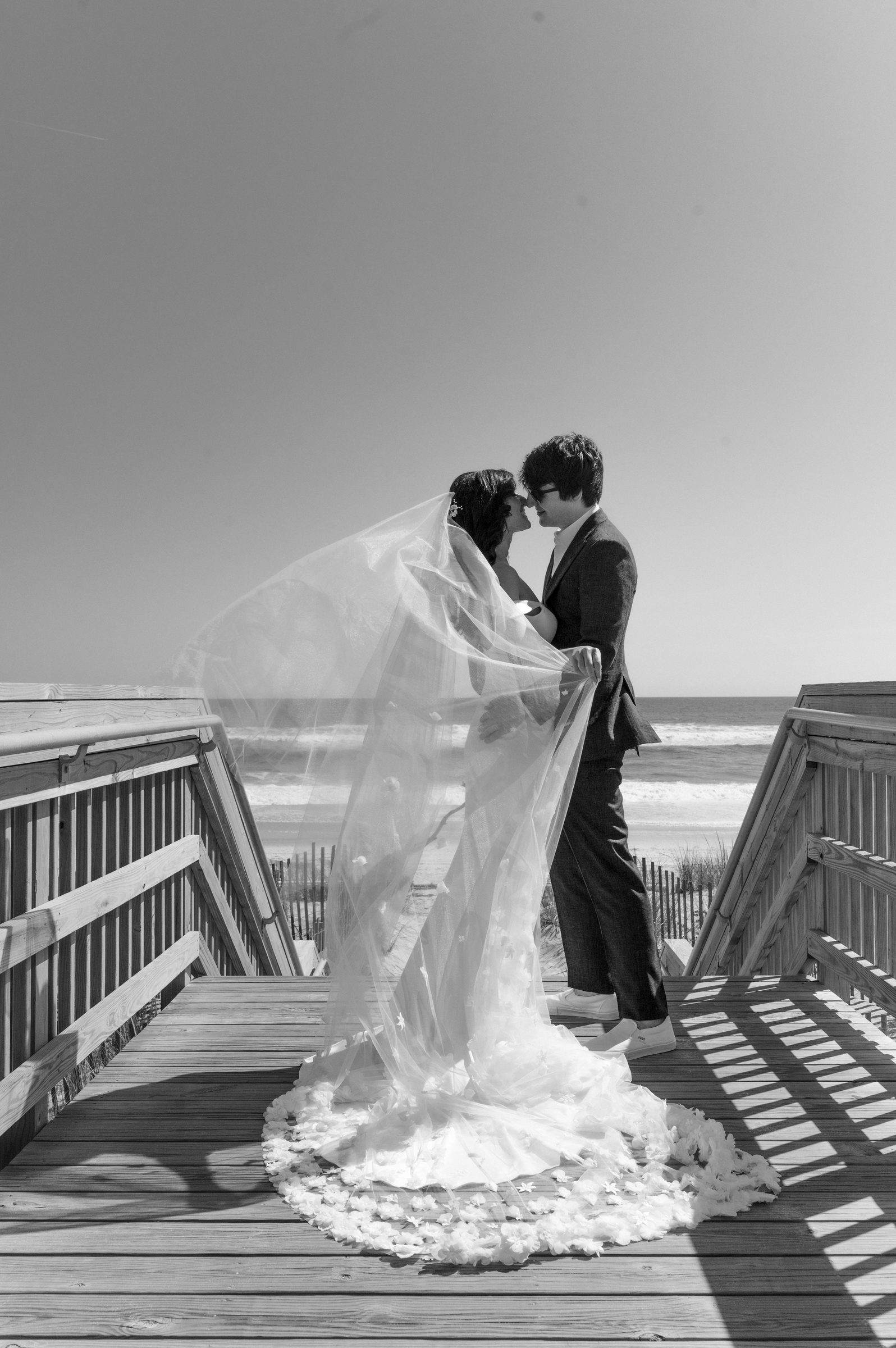 Black and white photo of a bride and groom embracing beneath a flowing veil on a beach boardwalk