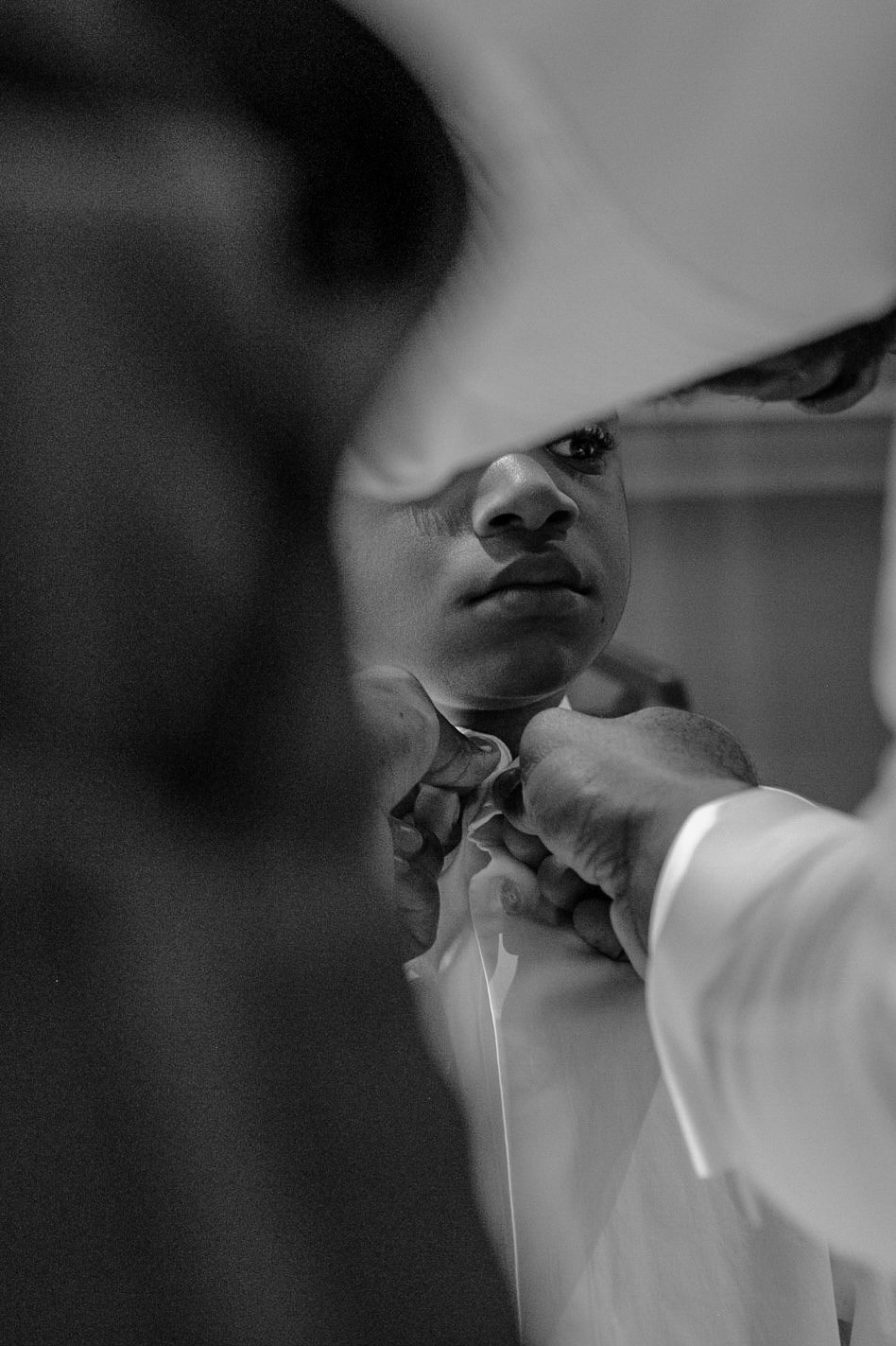 Black and white close-up of a young boy being buttoned into his shirt before the ceremony
