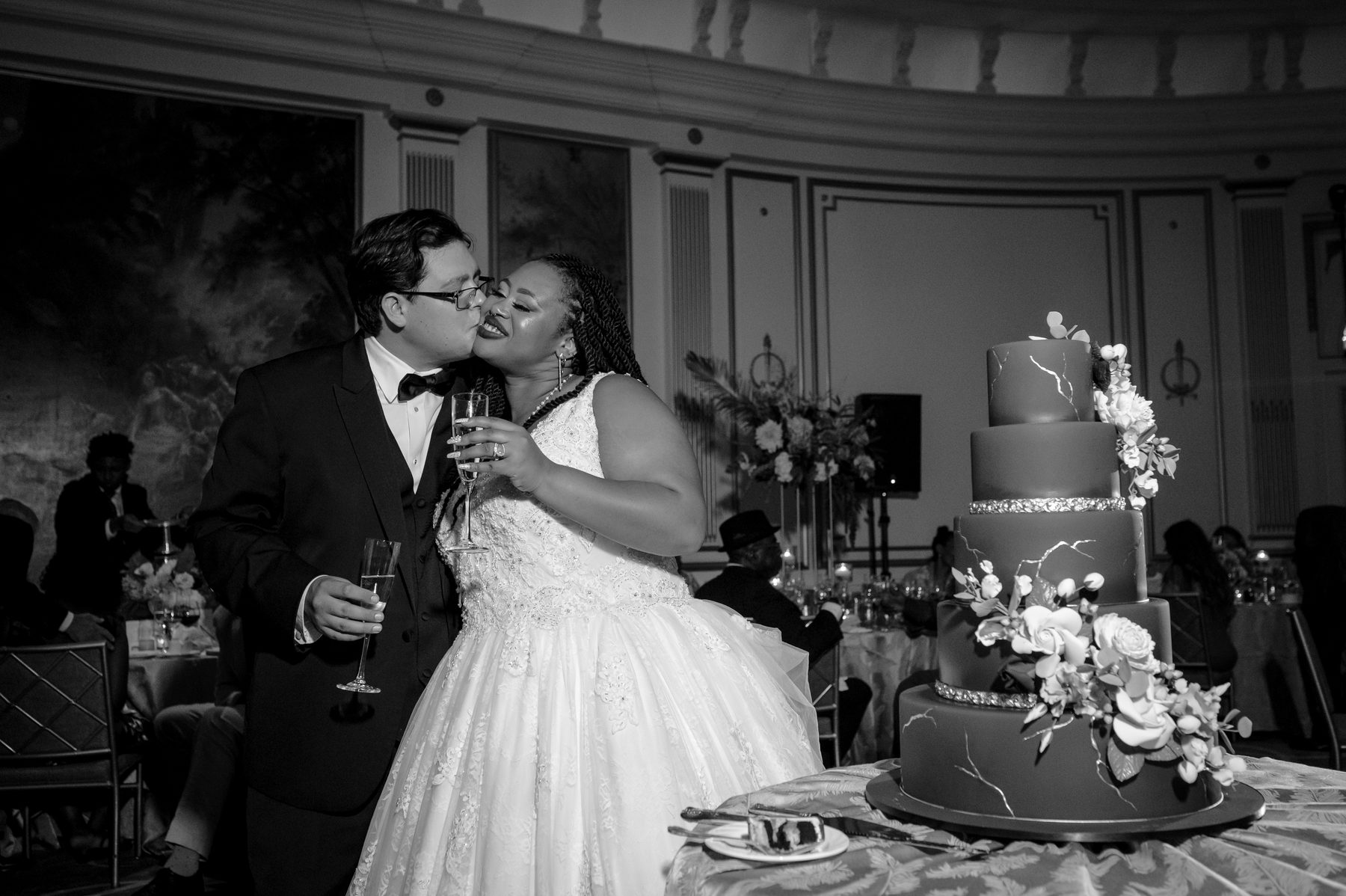 Black and white photo of a bride and groom kissing beside their wedding cake