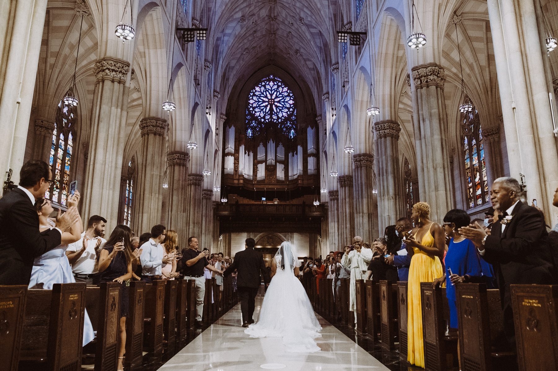 Bride and groom recessing down the aisle of a grand cathedral as guests applaud