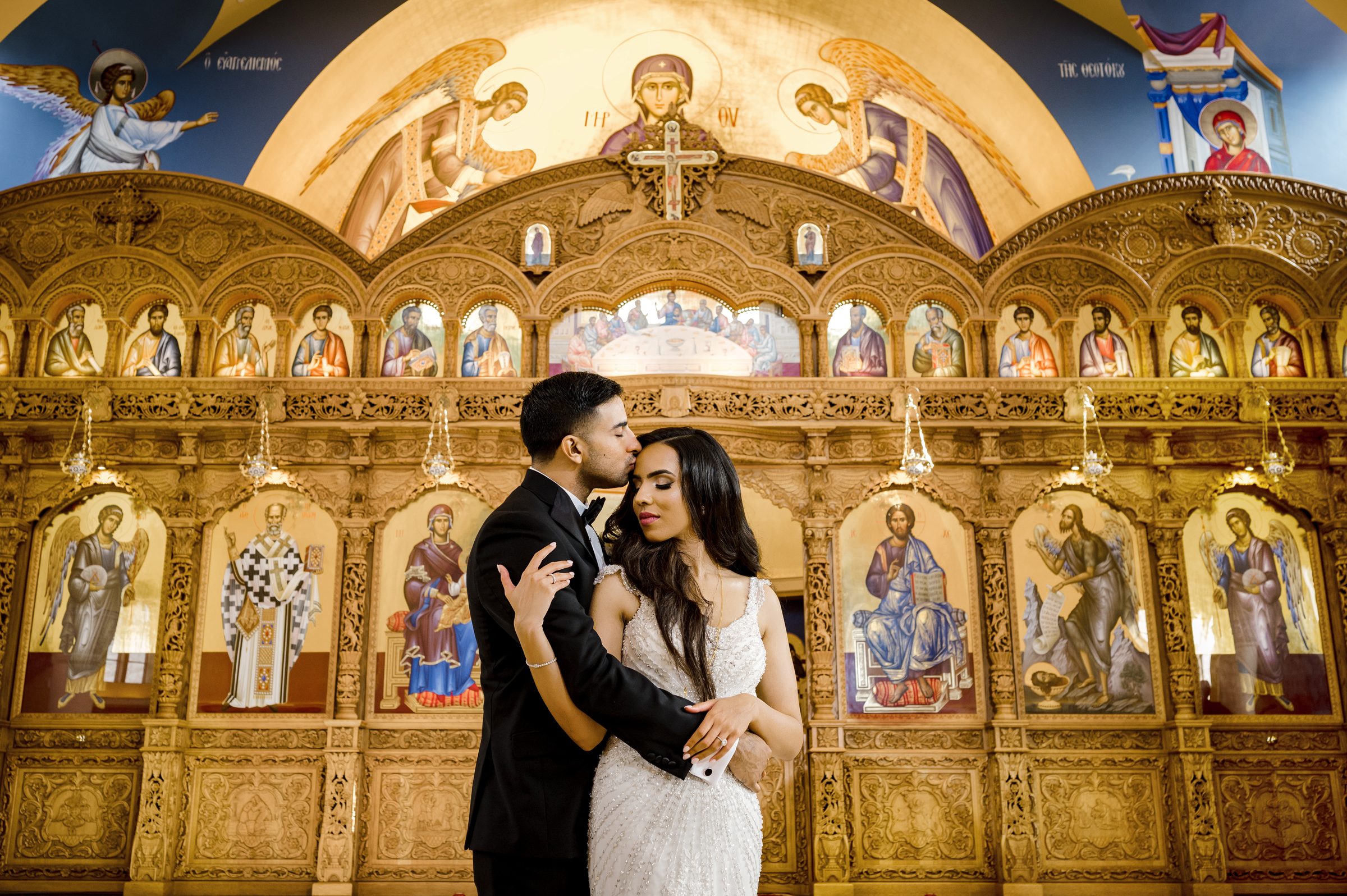 Groom kissing his bride's forehead in front of an ornate gold cathedral iconostasis