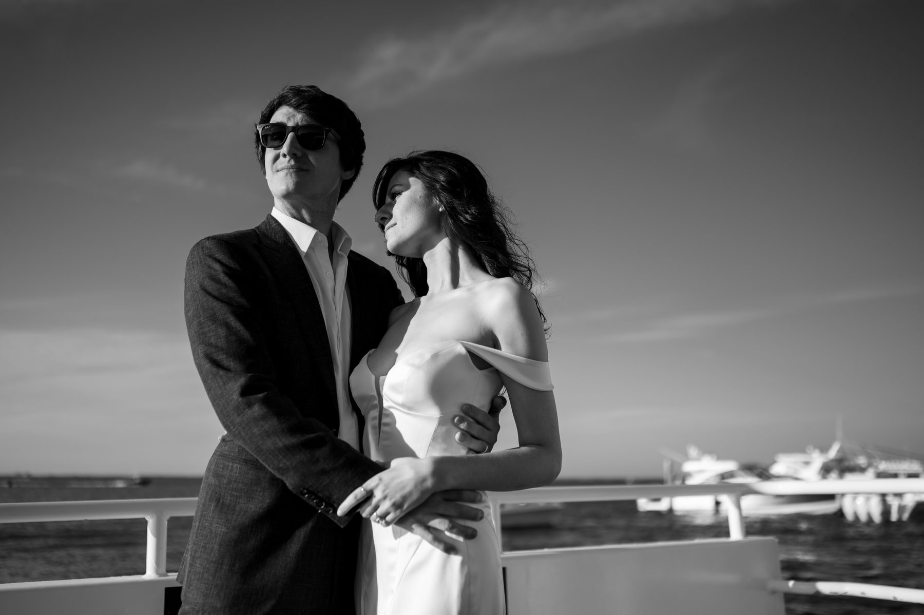 Black and white photo of a couple standing together on a boat deck with the marina behind them