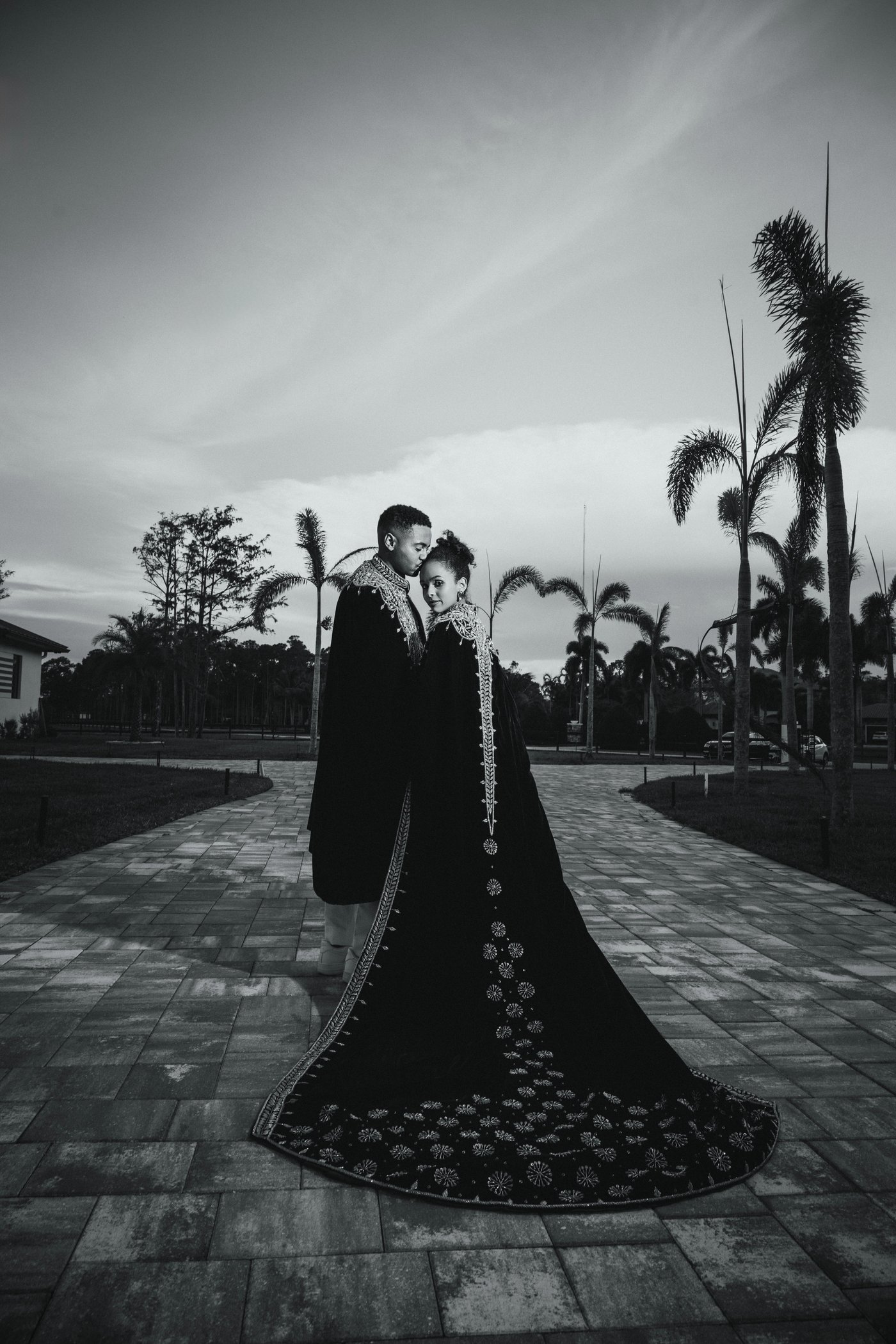 Black and white photo of a couple in dramatic couture attire standing on a path beneath tall palm trees