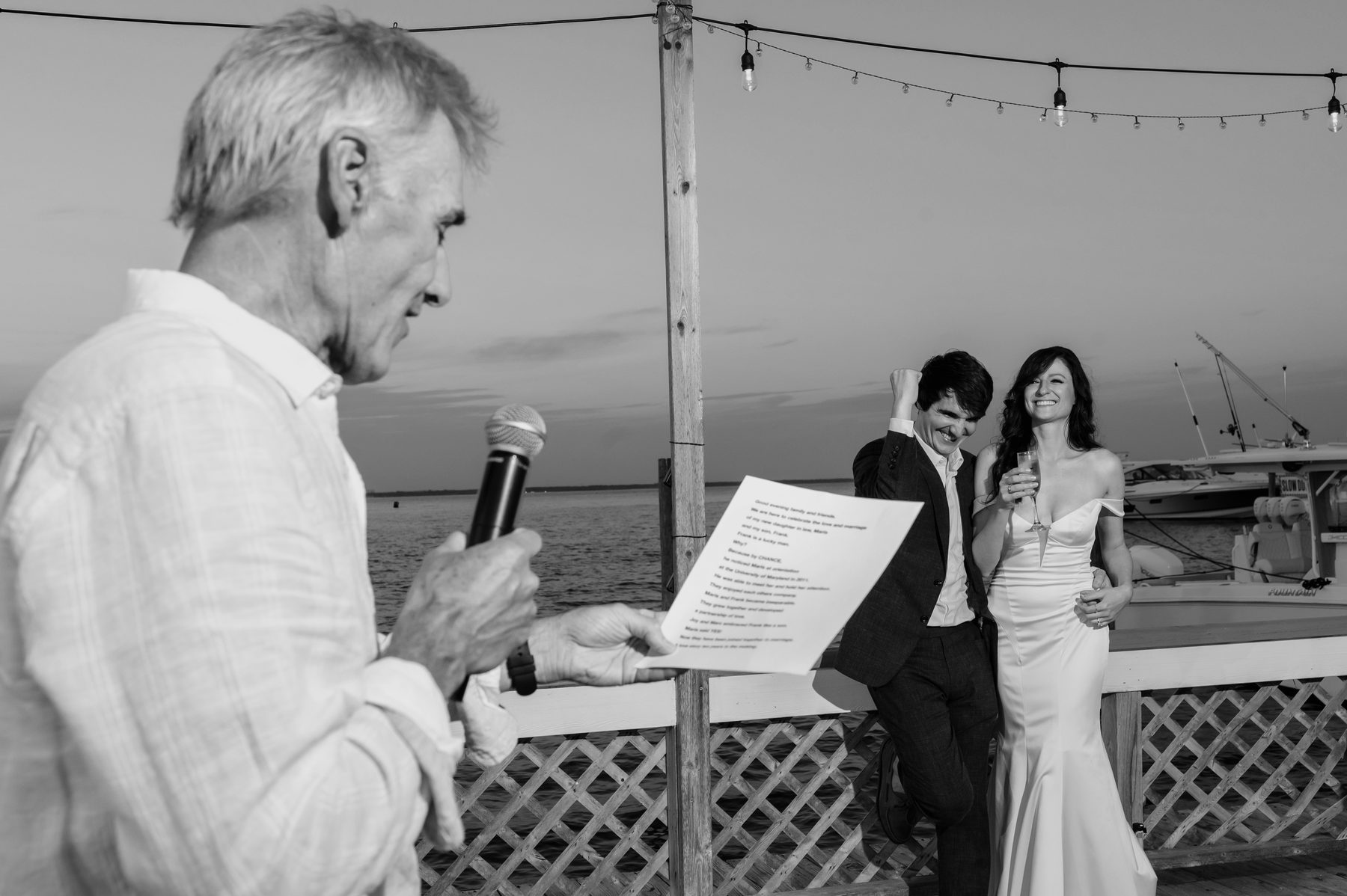 Black and white photo of a father giving a toast on a dock at sunset as the couple laughs