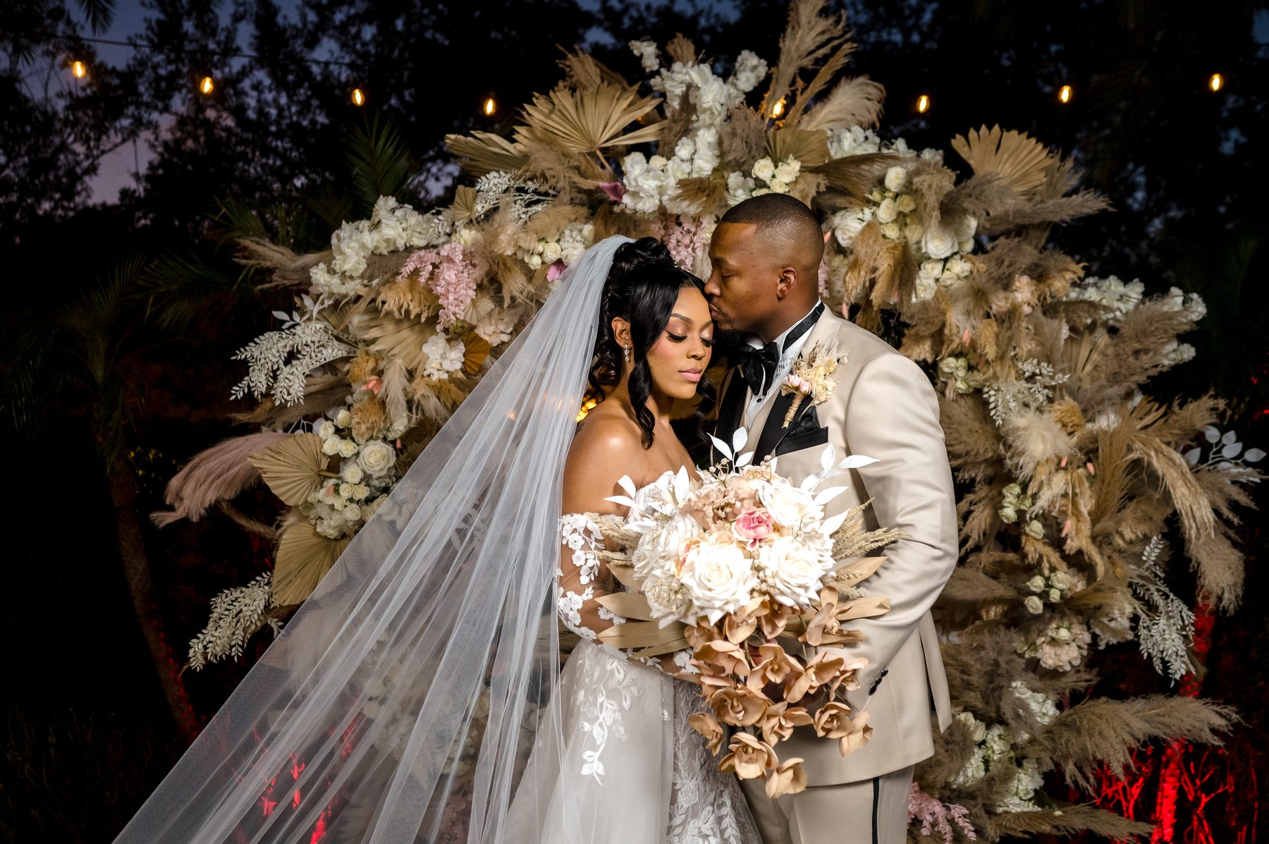 Groom kissing his bride's forehead in front of a dramatic dried floral installation at night