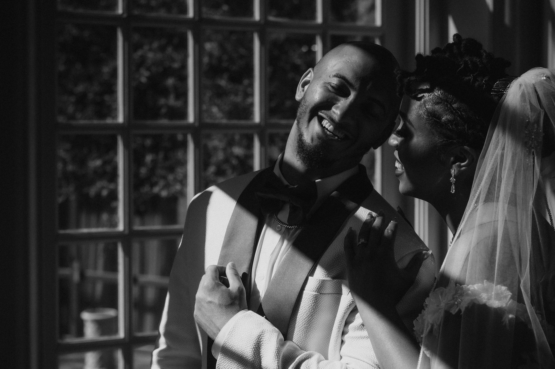 Black and white portrait of a bride and groom laughing together by a window