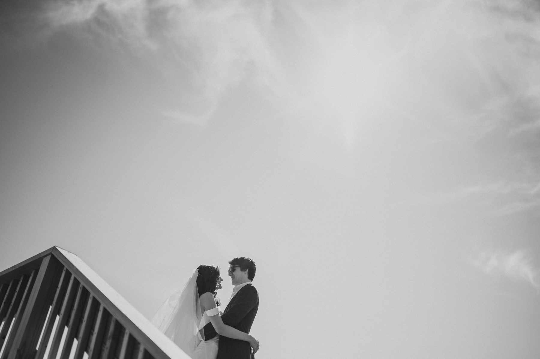 Black and white photo of a couple embracing beneath a peaked rooftop against a cloudy sky