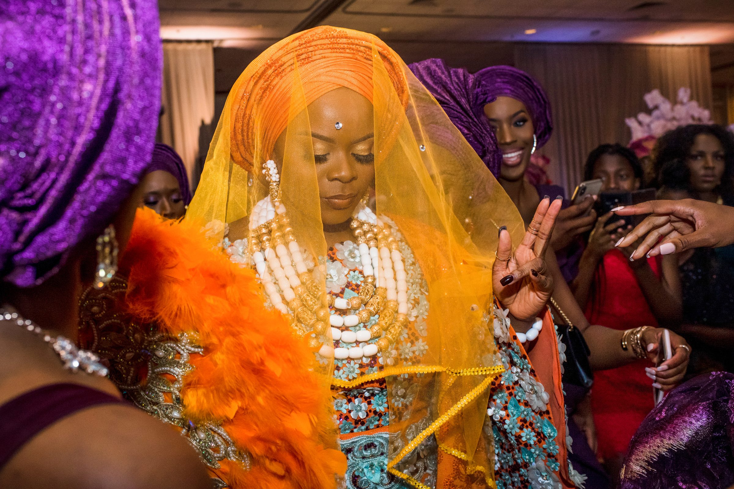 Bride in a golden yellow gele headwrap surrounded by guests capturing the moment