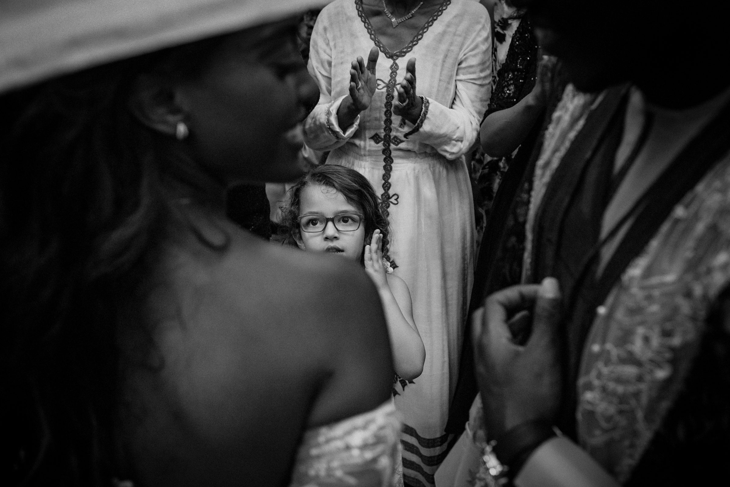 Black and white photo of a young girl in glasses watching quietly among the wedding guests