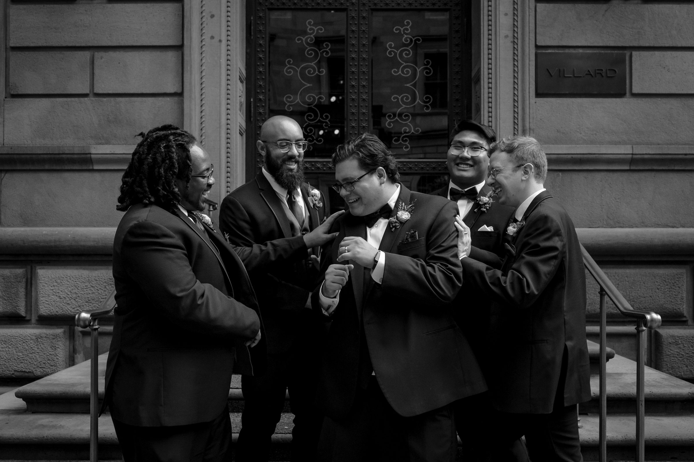 Black and white photo of five groomsmen laughing together outside a historic building