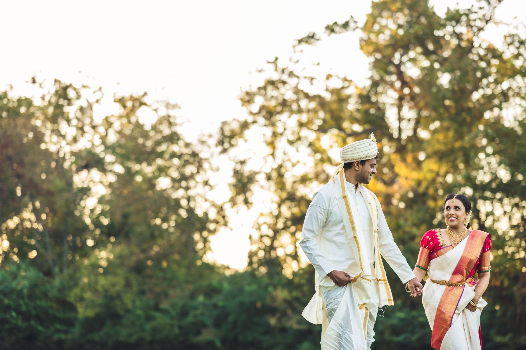 Groom in traditional cream attire and bride in a red and gold saree walking together outdoors