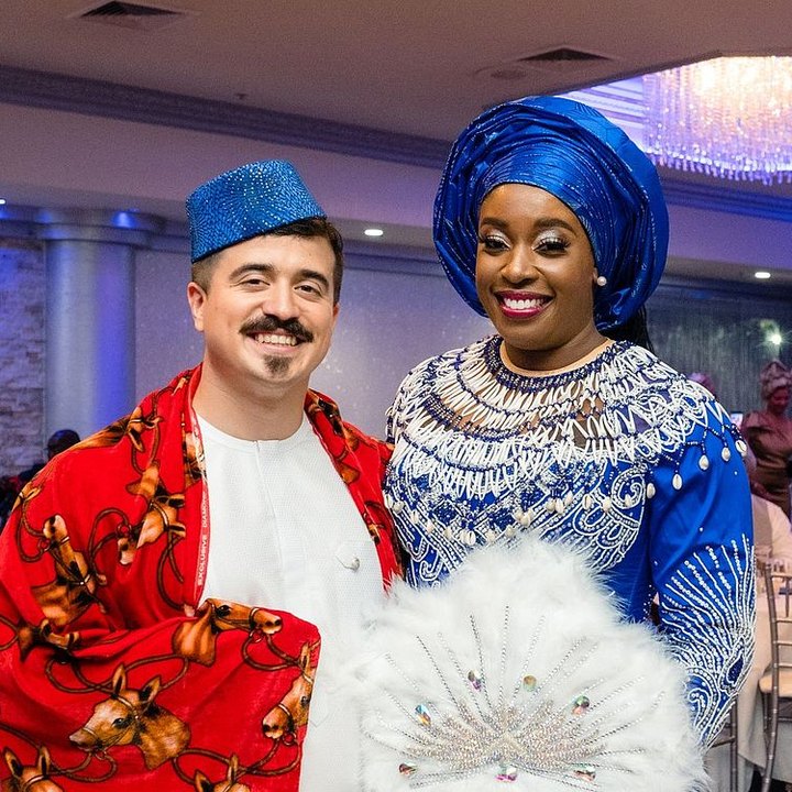 Tina and Chance in traditional wedding attire at their reception, her in blue with a white feather fan, him in a red patterned robe
