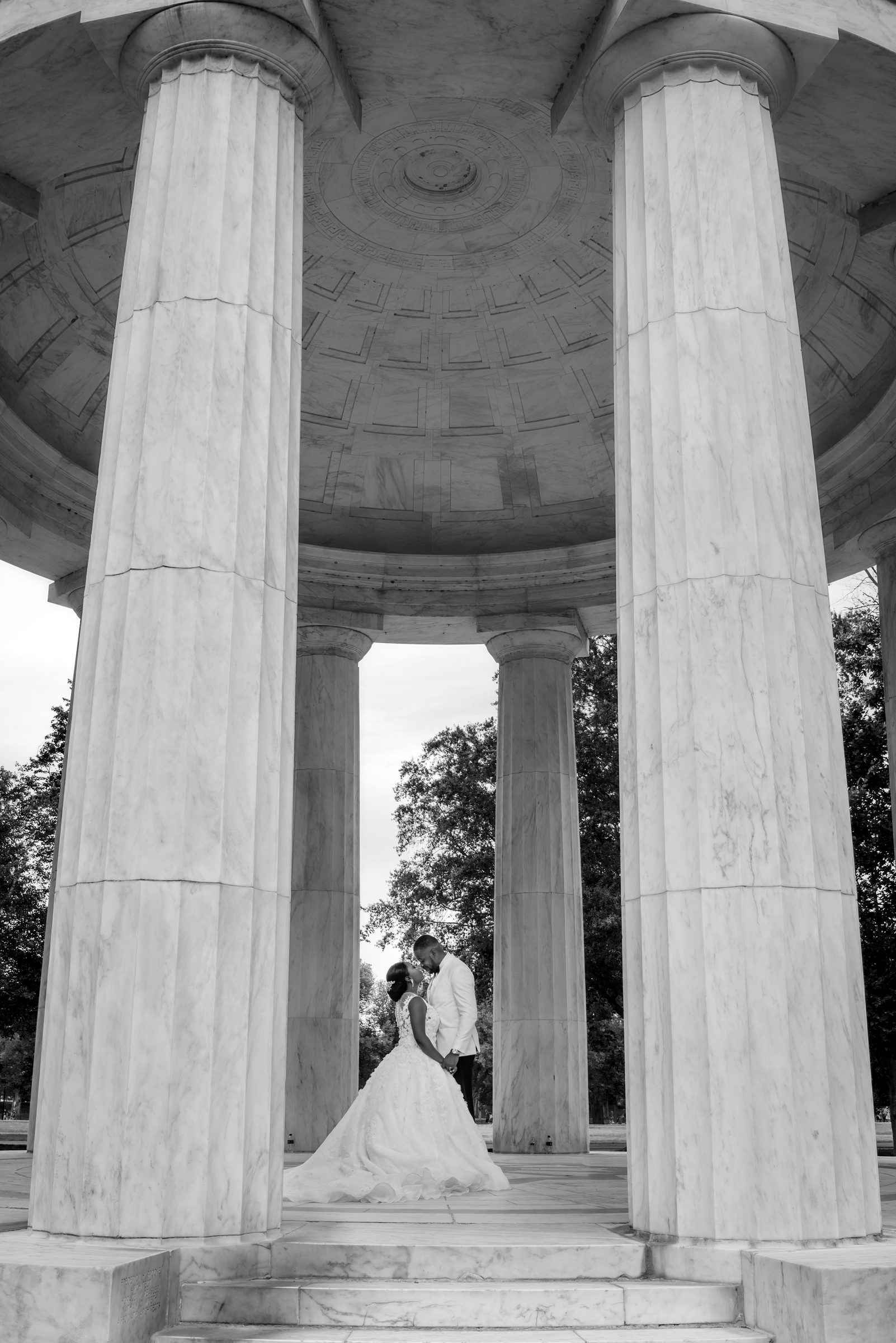 Black and white photo of a bride and groom sharing a kiss beneath the towering marble columns of a memorial rotunda