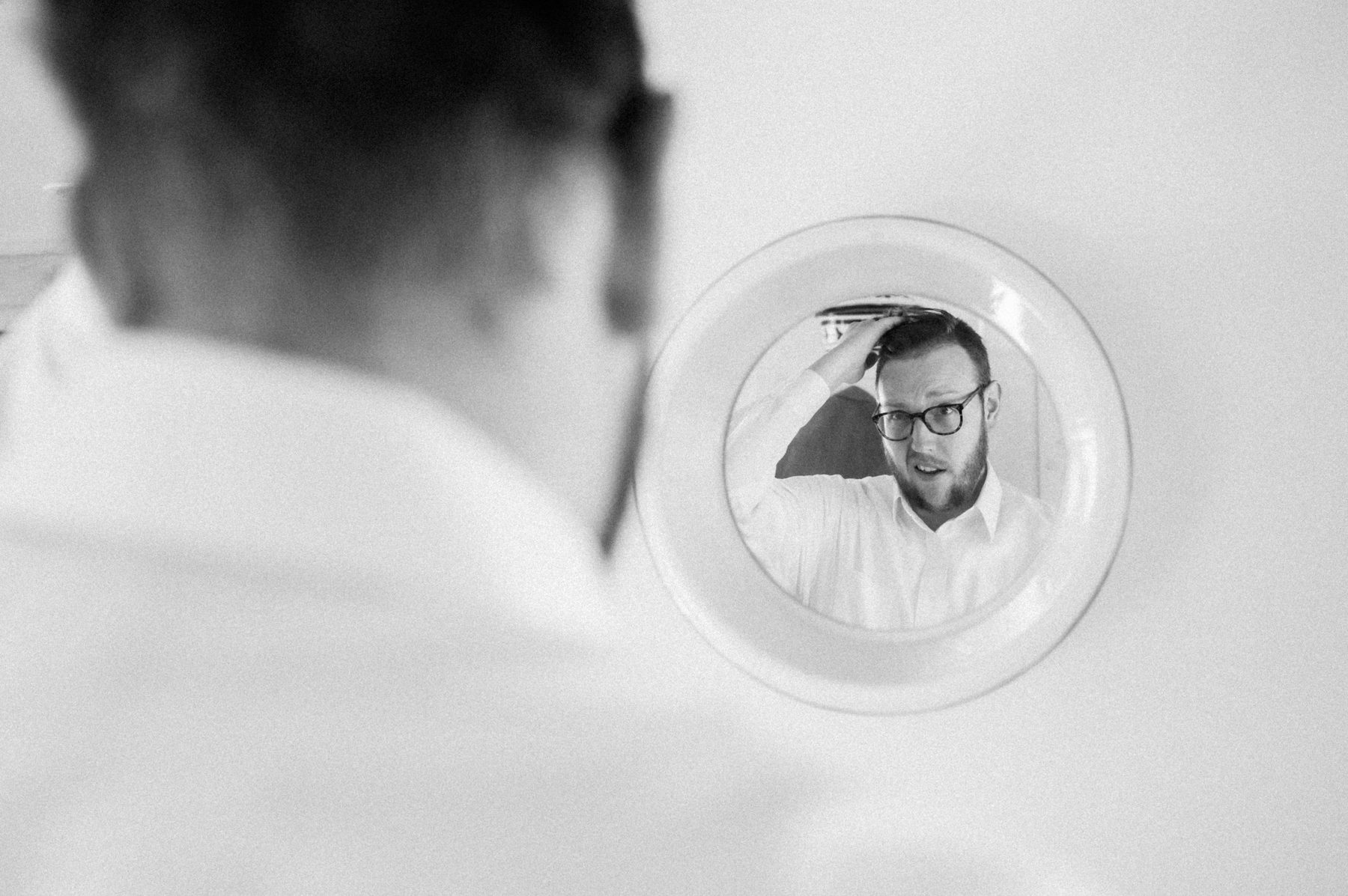 Black and white photo of a groom checking his reflection in a round mirror while getting ready