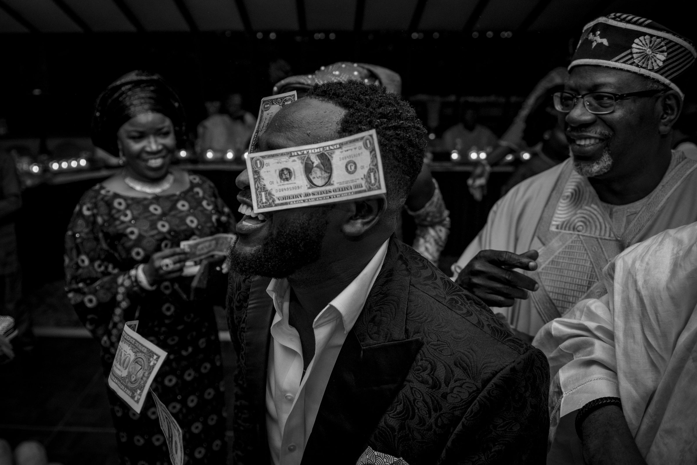 Black and white photo of a groom laughing with dollar bills covering his eyes during a money spray tradition