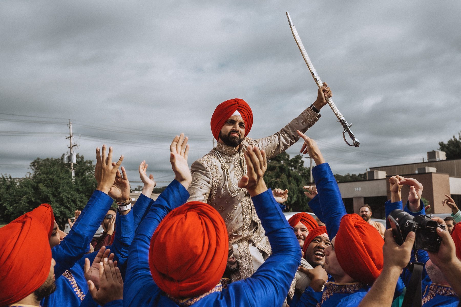 Sikh groom in a red turban raising a sword above the crowd carrying him on their shoulders